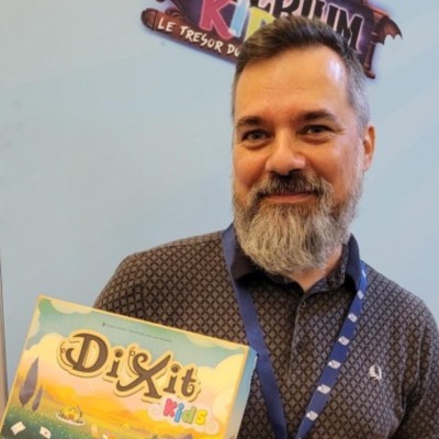 Un homme barbu, vêtu d'un polo foncé, sourit et tient une boîte du jeu de société « Dixit Kids » devant un fond bleu pâle, sur lequel on aperçoit partiellement du texte en haut.