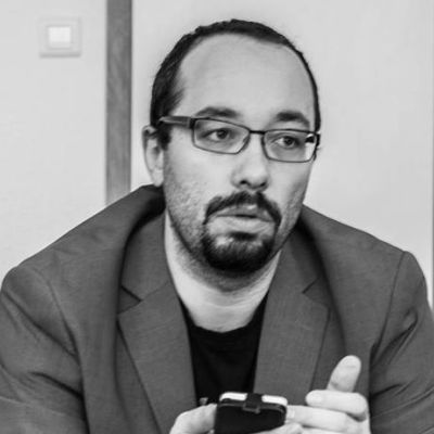 Un homme à lunettes et barbu, vêtu d'un blazer, a l'air pensif, un téléphone à la main. L'image est en noir et blanc.