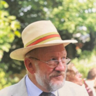 Un homme d'un certain âge, à la barbe grise et portant des lunettes, est vêtu d'un costume couleur crème, d'une chemise bleue et d'un chapeau de paille orné d'un ruban coloré. Il se tient debout à l'extérieur, avec de la verdure en arrière-plan. Une autre personne, légèrement floue, se trouve derrière lui.